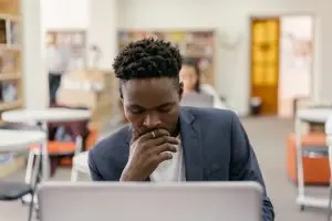 Male student looking at his computer in a classroom while he considers a college transfer.
